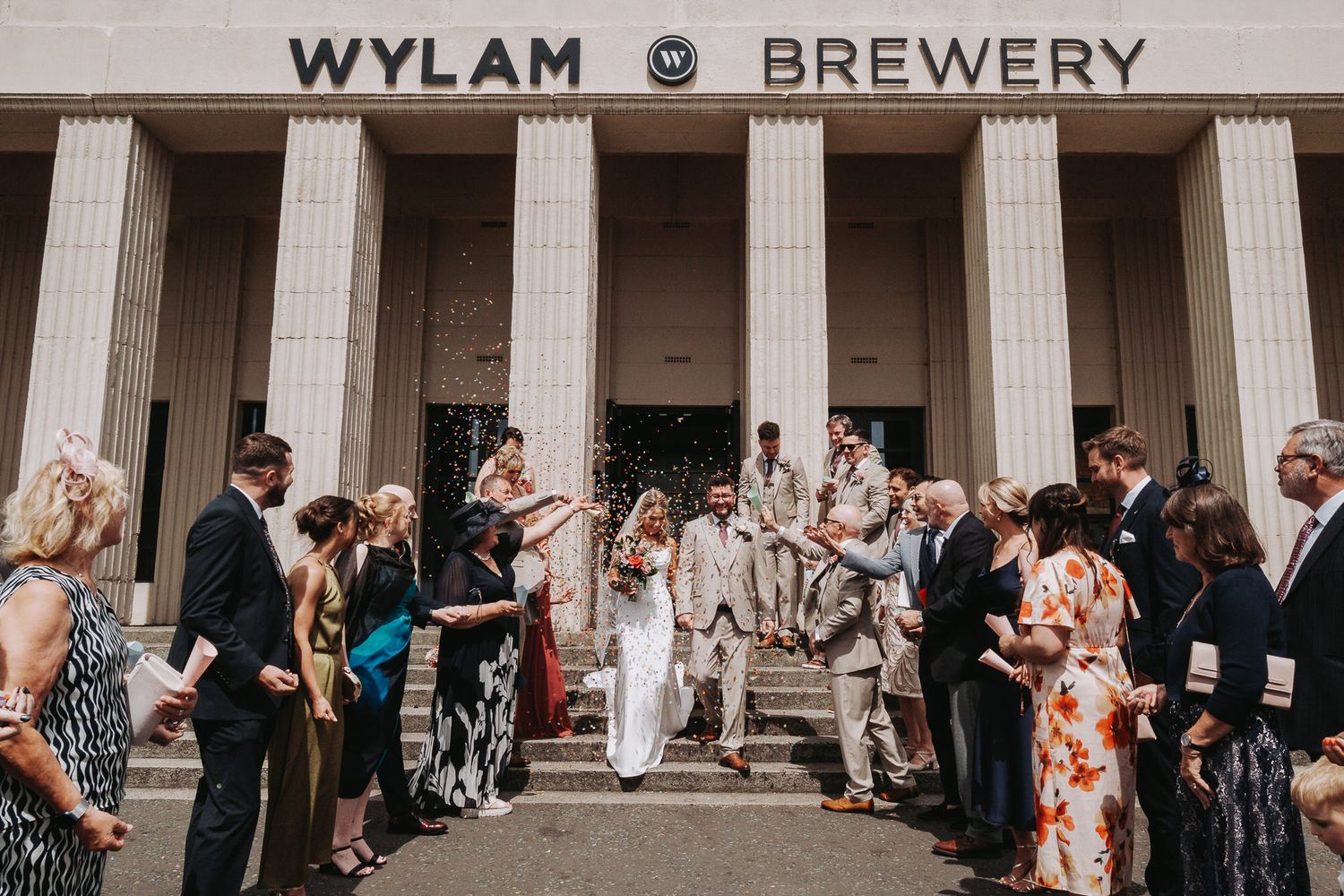 Newlyweds exit Wylam Brewery wedding venue surrounded by cheering guests throwing confetti on sunny steps.