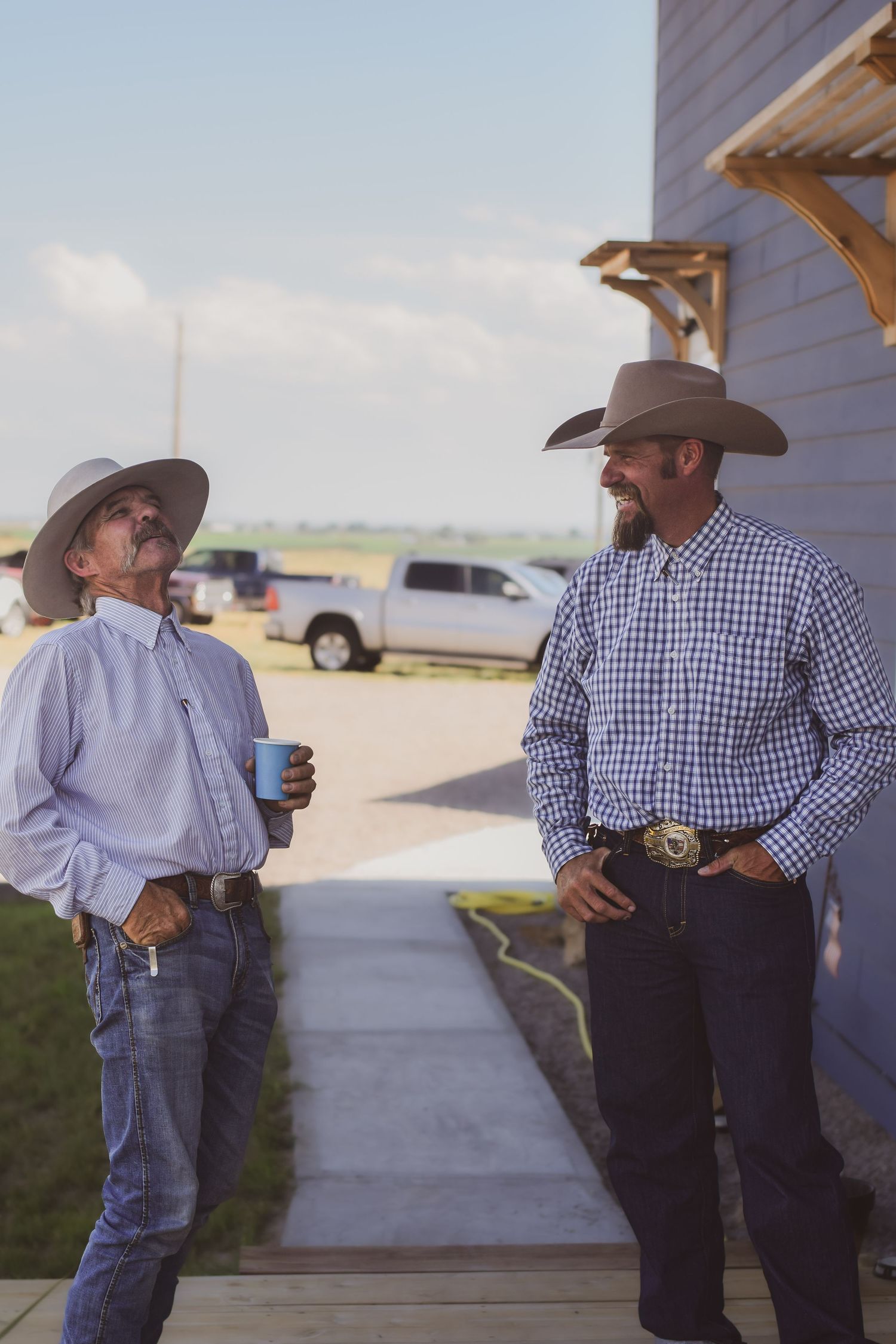 Two cowboys in hats and western shirts chat outside a ranch building on a sunny day.