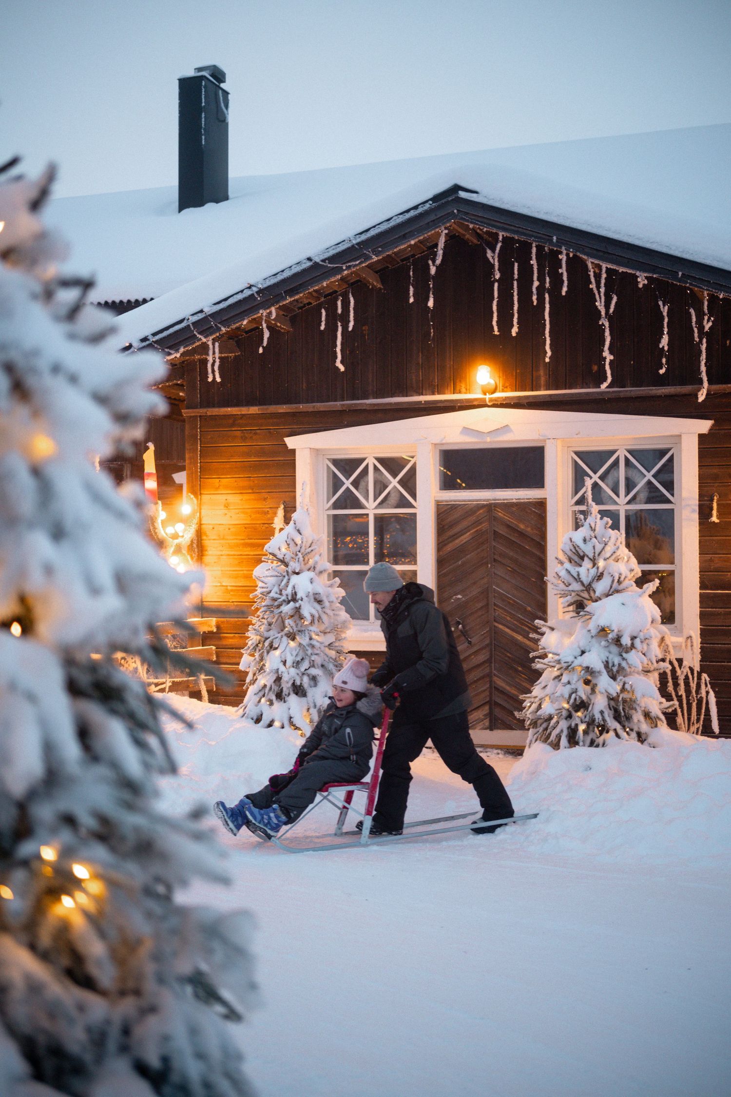 Person pushing a sled through snow past a cozy lit wooden cabin with icicles and decorated Christmas trees in winter.