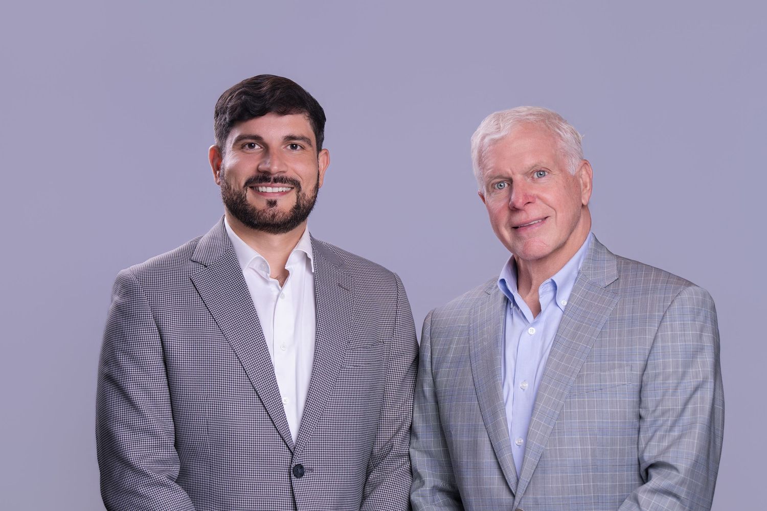 Two professional men in suits smiling, posing together against a gray background for a formal portrait.