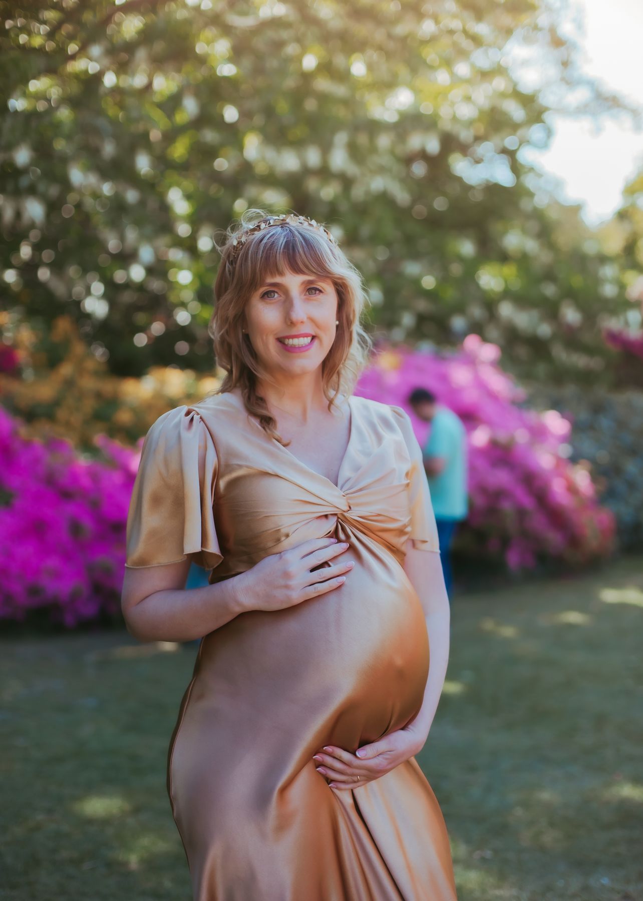 Pregnant woman in gold satin dress and floral crown poses outdoors among pink flowers. during a maternity photoshoot in Bexley, London