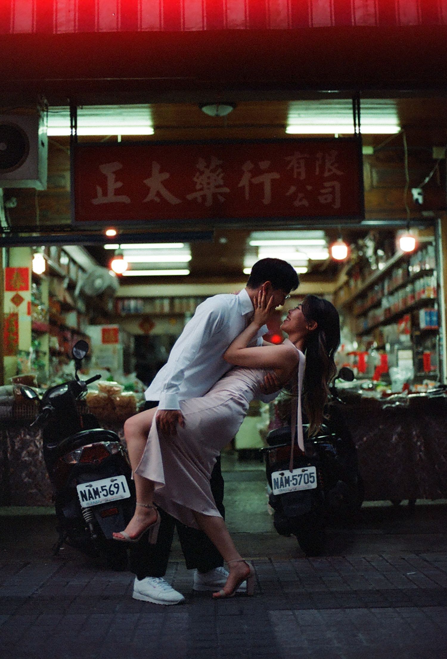 A couple shares a romantic kiss in front of a traditional Chinese pharmacy shop at night, with motorcycles parked nearby.