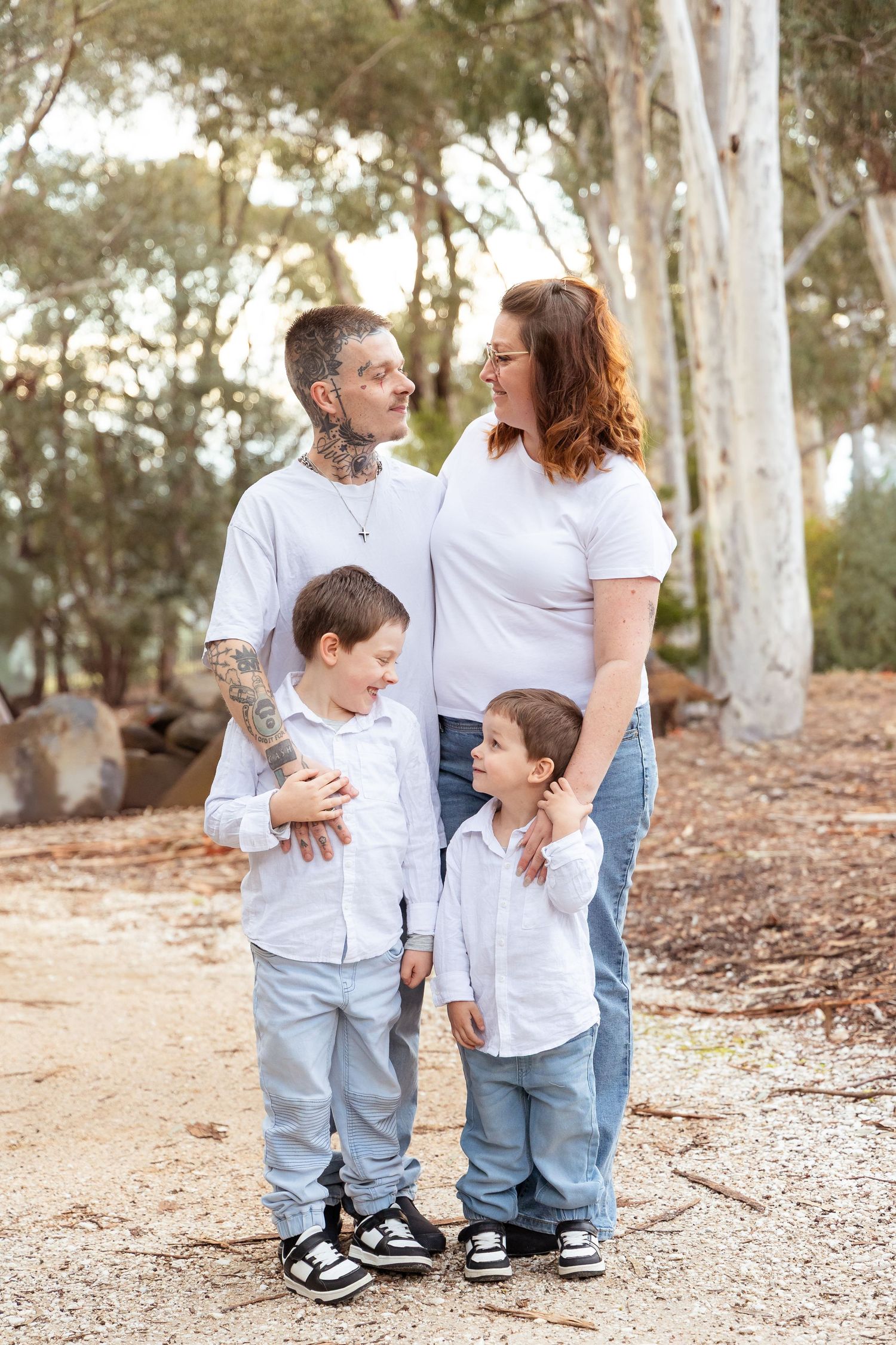 Family of four in white shirts and jeans posing outdoors among eucalyptus trees, parents with two young boys smiling.