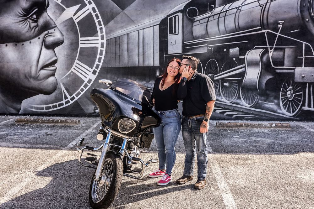 Couple posing in front of a large mural featuring a train and clock, with a motorcycle parked nearby in a parking lot.