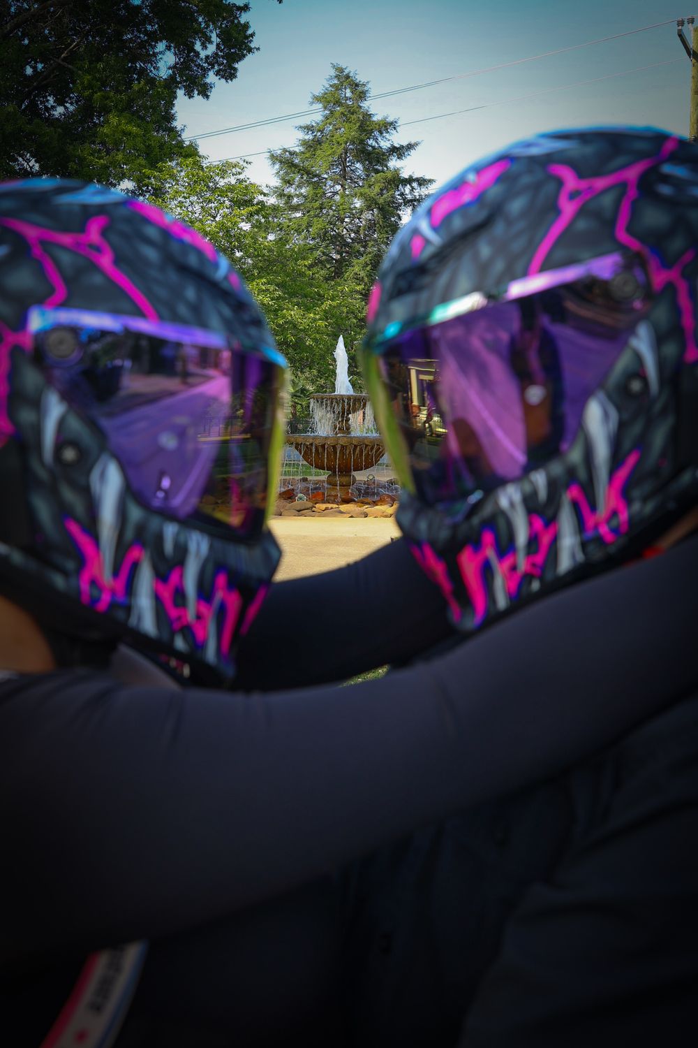 Two matching black and pink monster motorcycle helmets worn by riders facing a decorative fountain in a tree-lined park.