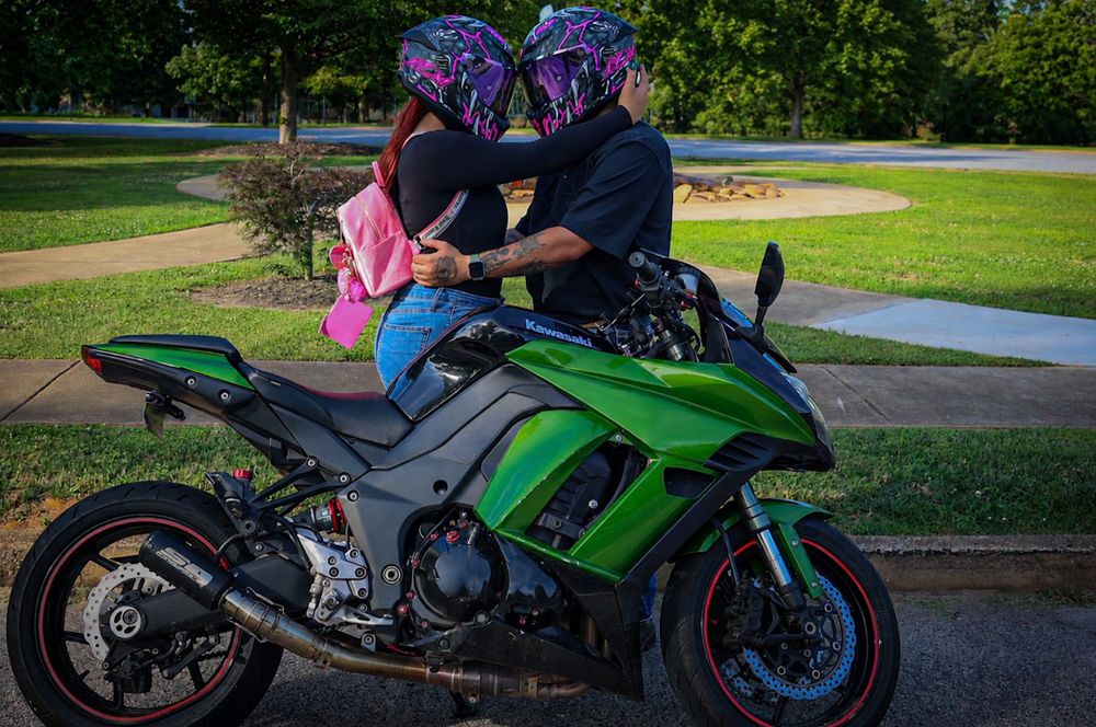 Two riders in colorful helmets share a romantic moment beside a green Kawasaki sport motorcycle parked outdoors.