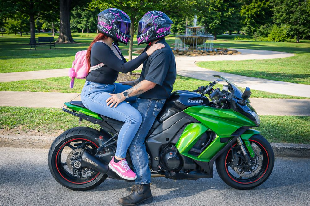 Couple wearing colorful helmets sitting on a green Kawasaki sport motorcycle in a sunny park.