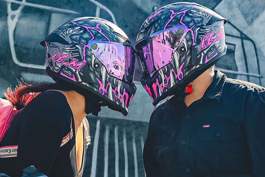 Two people wearing matching pink and black graffiti-style motorcycle helmets sharing a kiss outdoors.