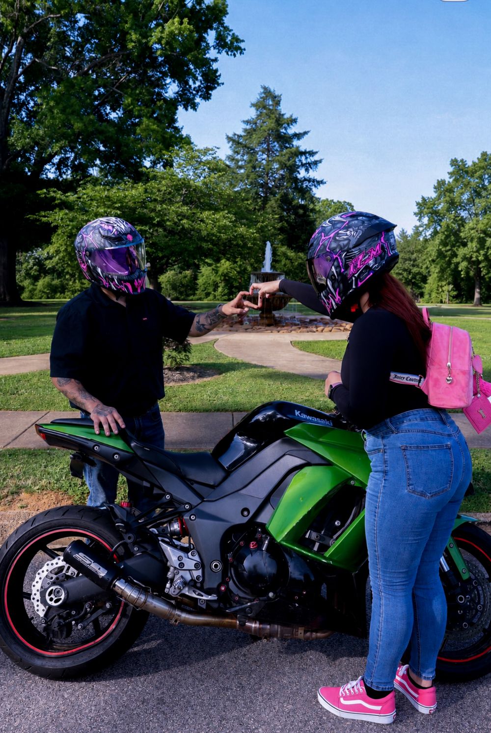 Two riders in matching pink helmets toast drinks beside a green Kawasaki sport bike in a scenic park.