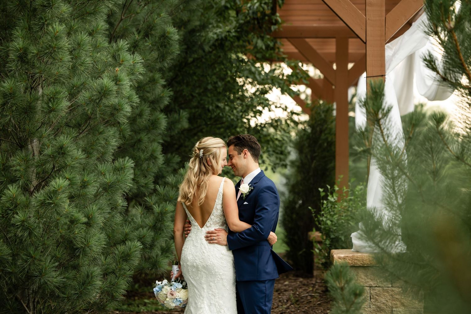Bride and groom share a romantic moment surrounded by lush evergreen trees near a wooden pergola on their wedding day.