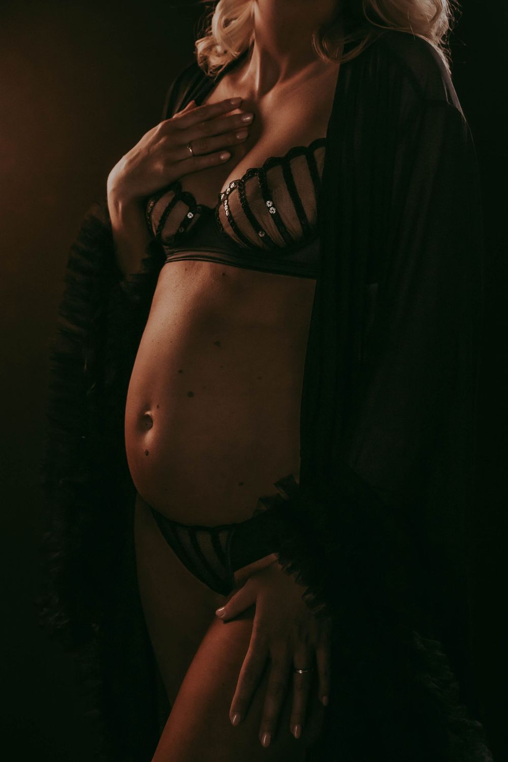Pregnant woman in black lingerie and robe posing elegantly in a dark, moody studio setting.