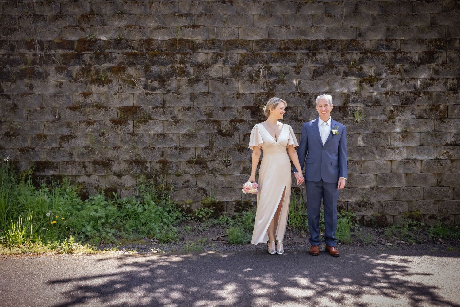 Bride in elegant champagne gown and groom in blue suit standing together before a rustic stone wall outdoors.