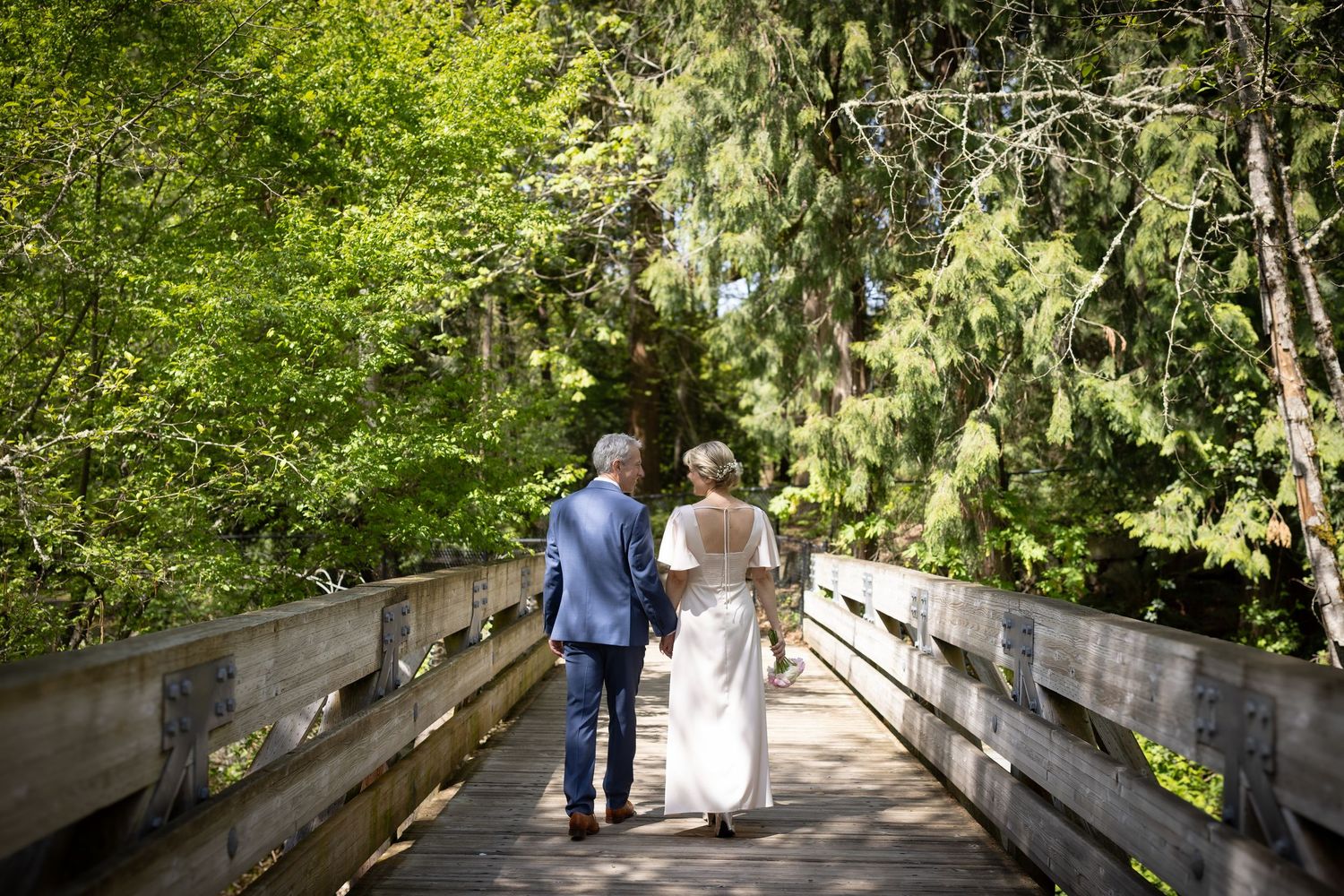 Couple in wedding attire walking hand-in-hand on a wooden bridge surrounded by lush green trees.