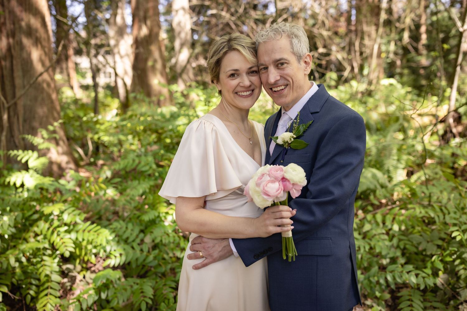 Smiling bride and groom pose together in a lush forest setting, bride holding pink bouquet, groom in navy suit.