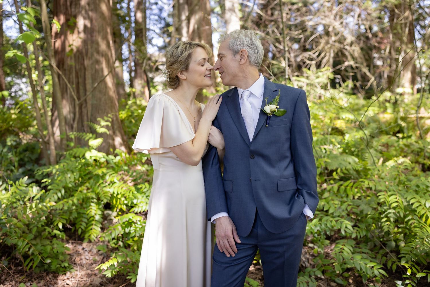A couple in wedding attire shares a romantic moment surrounded by lush green ferns and tall trees in a forest setting.