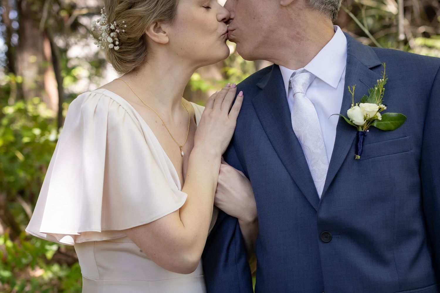 Bride and groom sharing a romantic kiss outdoors, wearing wedding attire with a white boutonniere and elegant necklace.