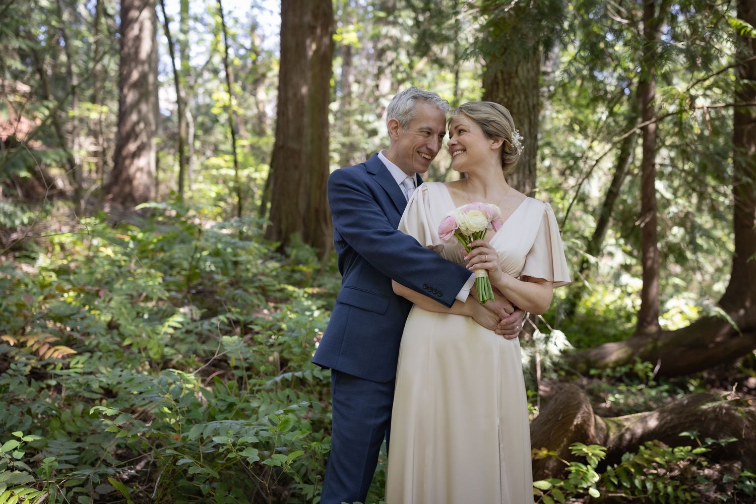 Couple in wedding attire embracing in a lush forest, bride holding pink bouquet in elegant white dress.