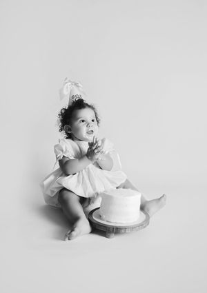 Black and white photo of a baby girl in a white dress and bow, sitting beside a small cake for her first birthday.
