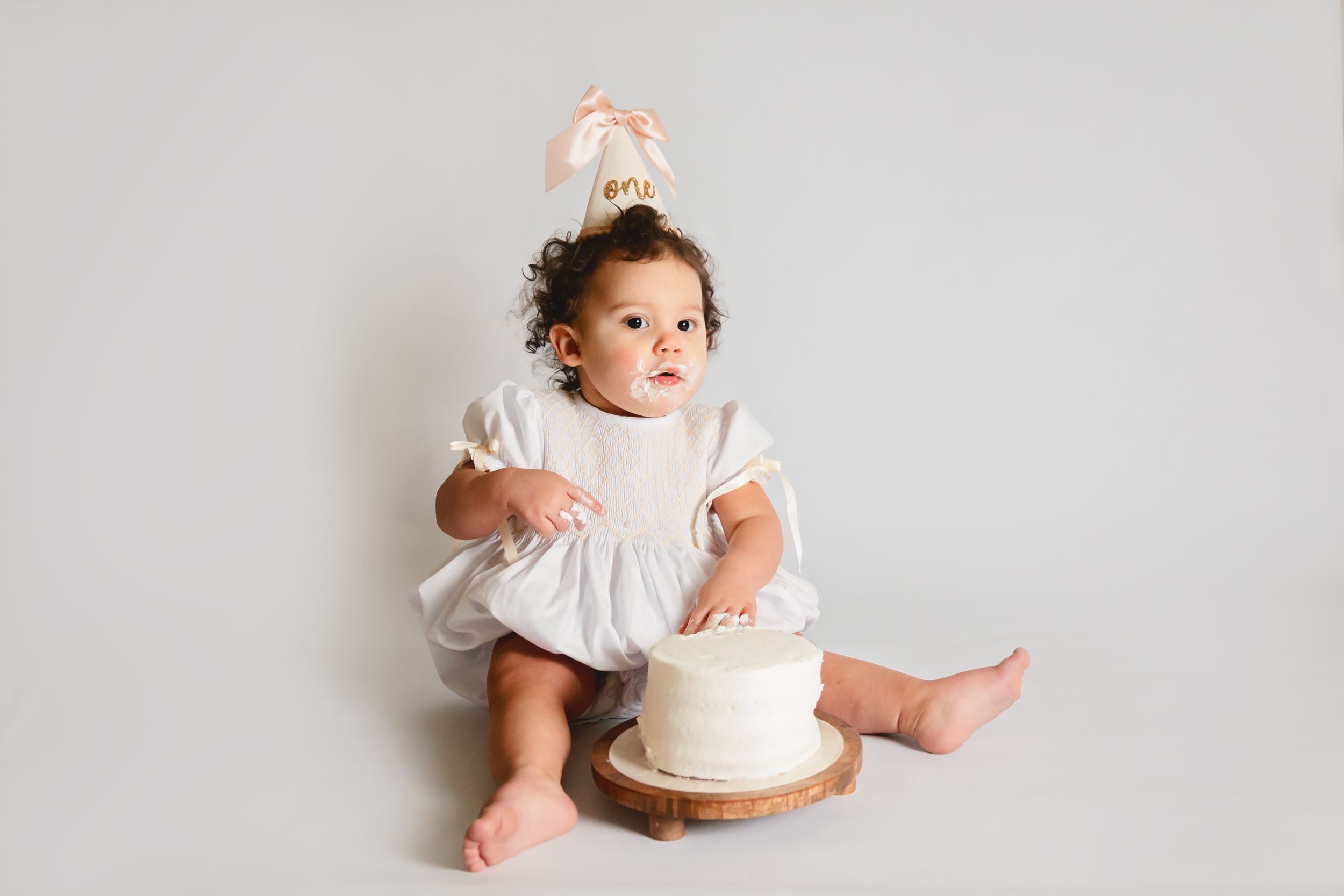 Baby girl in white dress with bow headband sits beside a white first birthday smash cake on a wooden stand.
