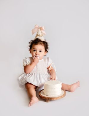 Baby girl in white dress and gold \u2018one\u2019 party hat smashing her first birthday cake on a wooden board.