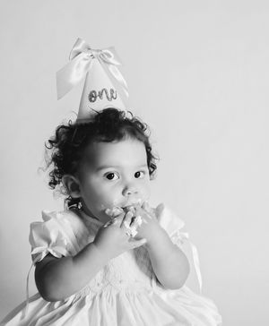 Black and white photo of a baby girl in white dress wearing a first birthday party hat eating cake.