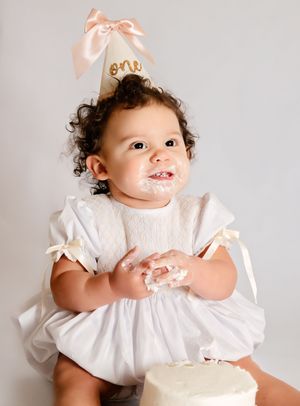 Adorable baby girl in white dress and pink \u2018one\u2019 party hat enjoying first birthday cake smash.