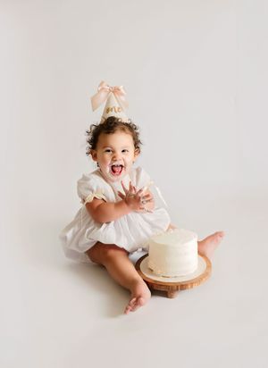 Baby girl in white romper and pink party hat laughing during first birthday cake smash photo session.