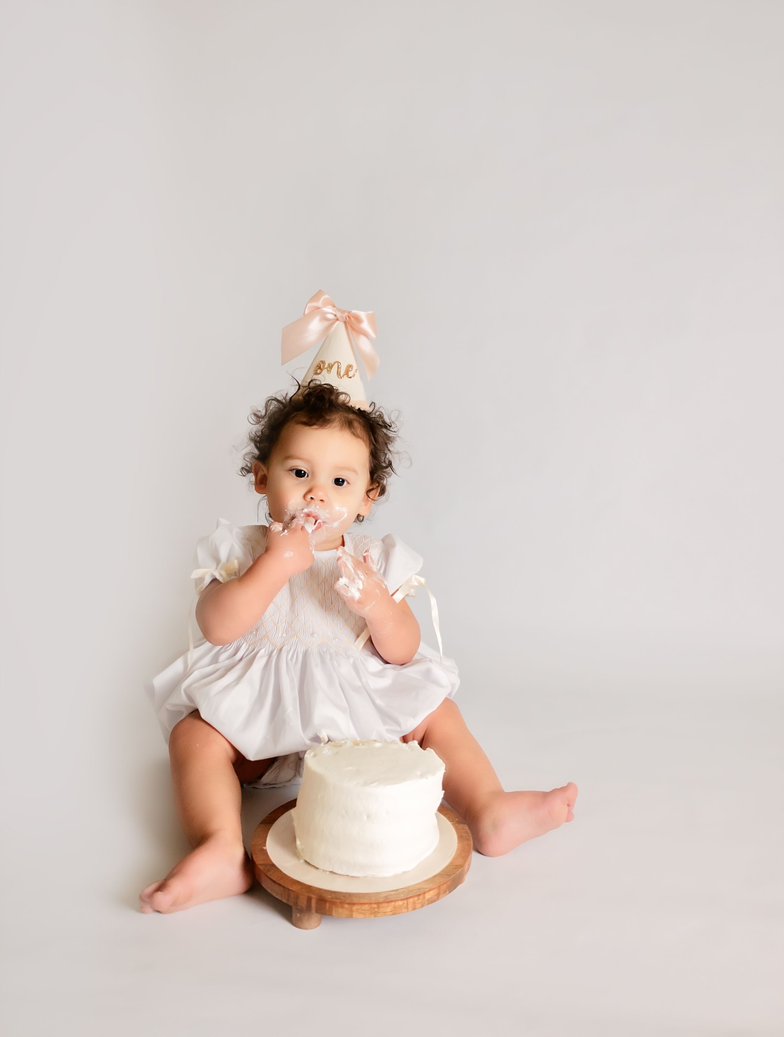 Baby girl in white dress and pink party hat eating cake during a first birthday cake smash photo session.