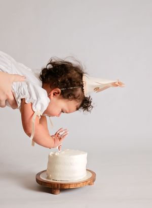 A toddler in a white dress leans forward to taste a white frosted smash cake on a wooden stand.
