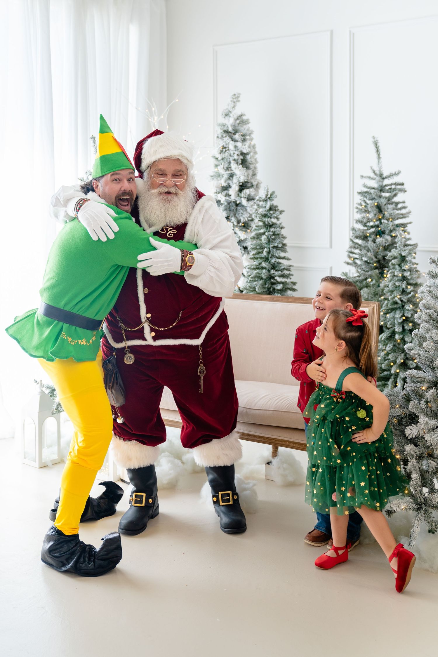 Family posing with Santa and an elf costume in front of flocked Christmas trees in a festive holiday photo session.