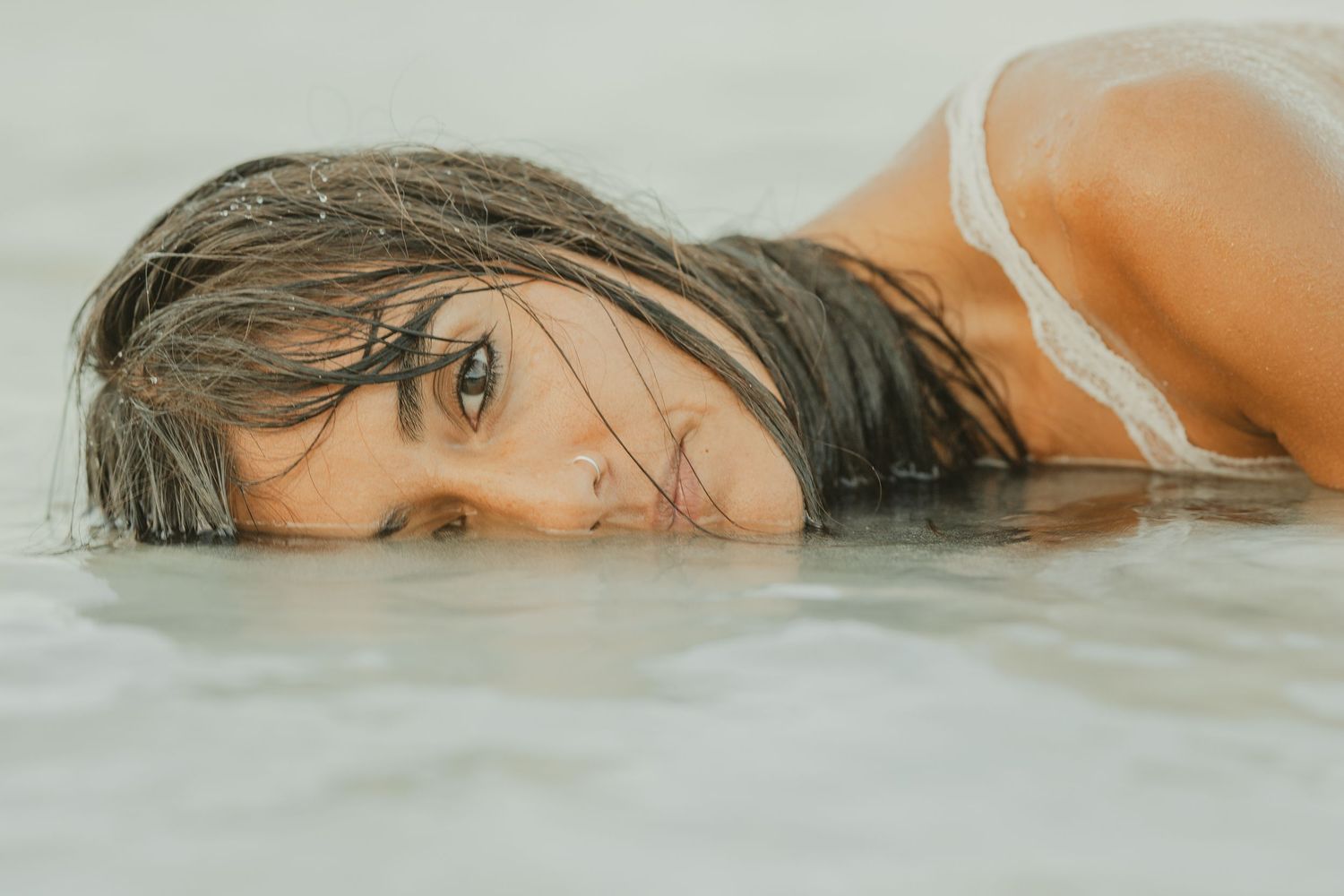 Young woman with wet hair and green eyes lies in shallow water, looking directly at the camera.