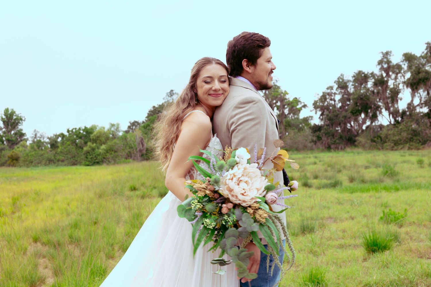 Bride in white gown hugs groom from behind holding bouquet in a lush green meadow on their wedding day.
