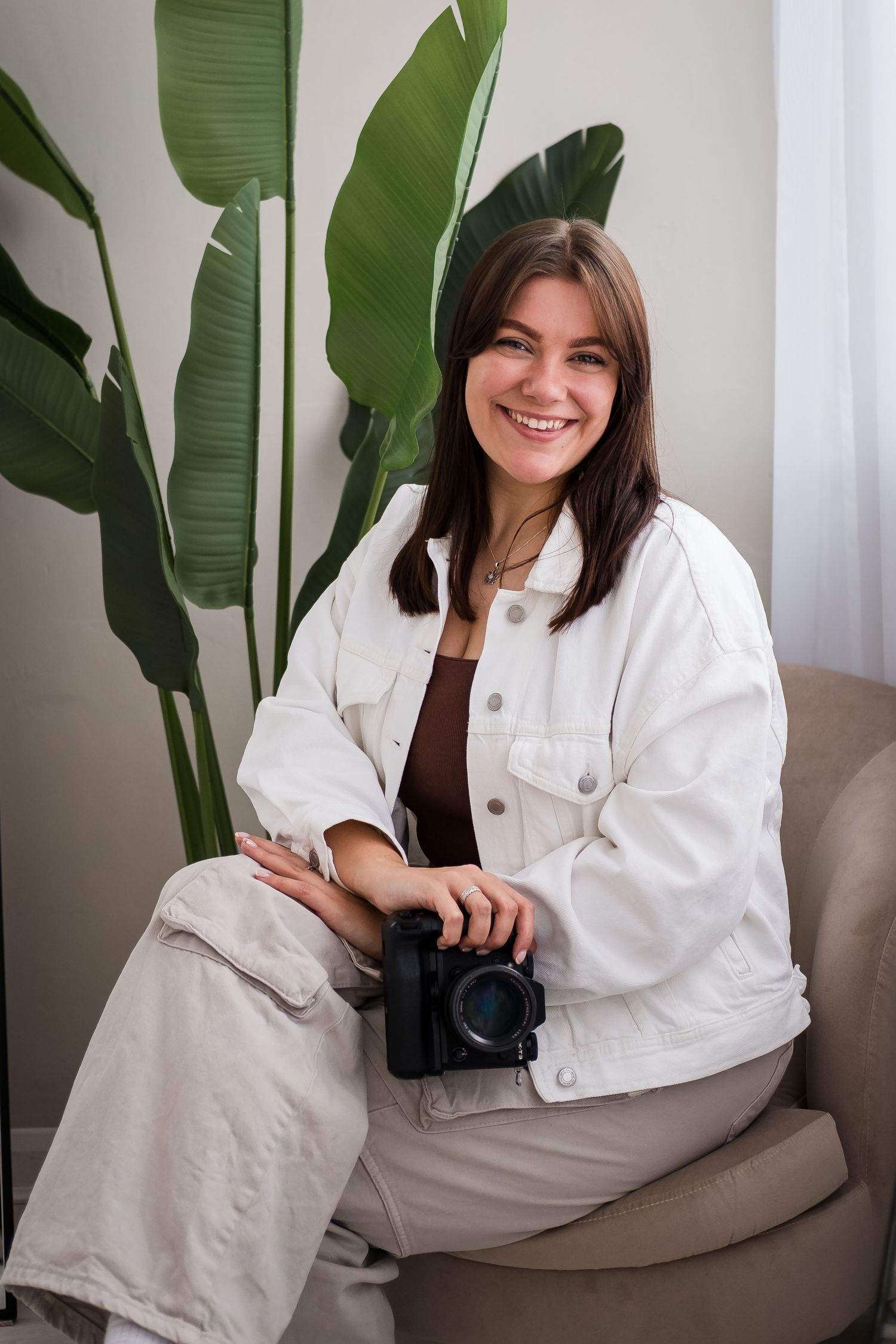 Smiling young woman in white denim jacket holding a camera, seated in a chair with a tropical plant backdrop.