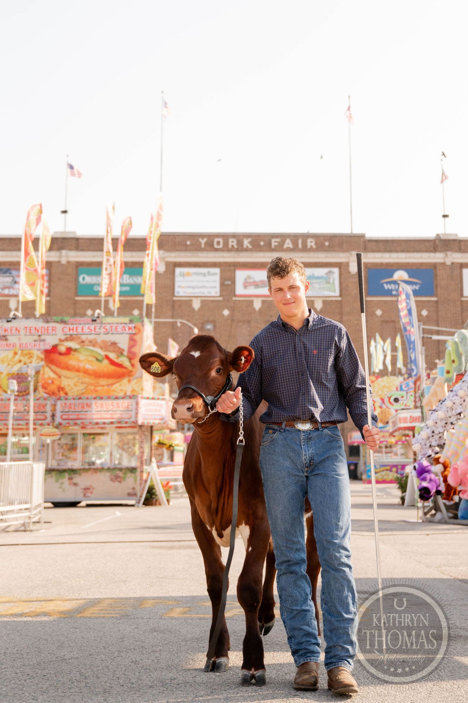Man stands with a brown cow and pink pig at a county fair with a Ferris wheel backdrop.