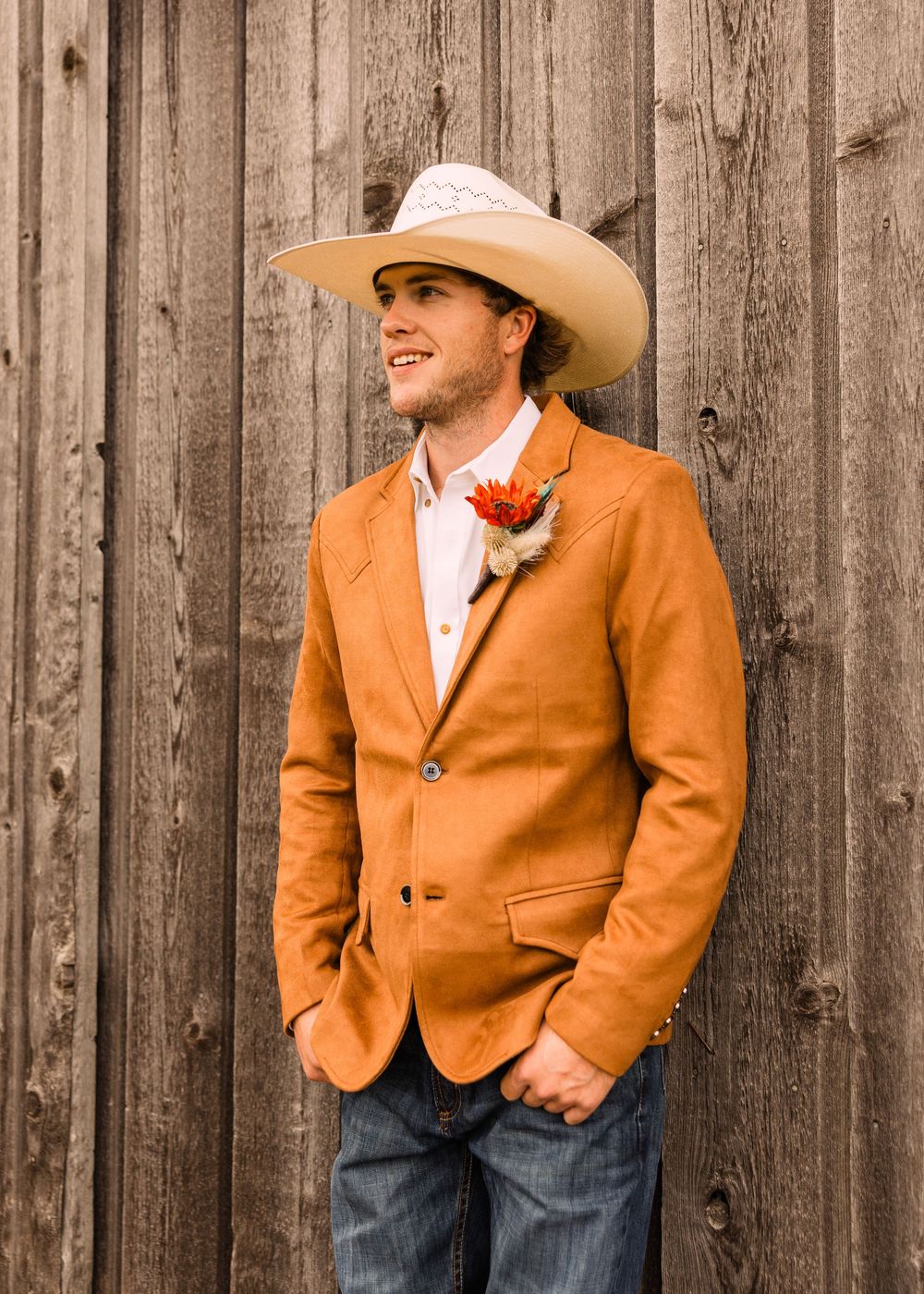 Smiling groom in a cowboy hat, orange suede blazer and jeans wears a floral boutonniere while leaning on a weathered barn wall.