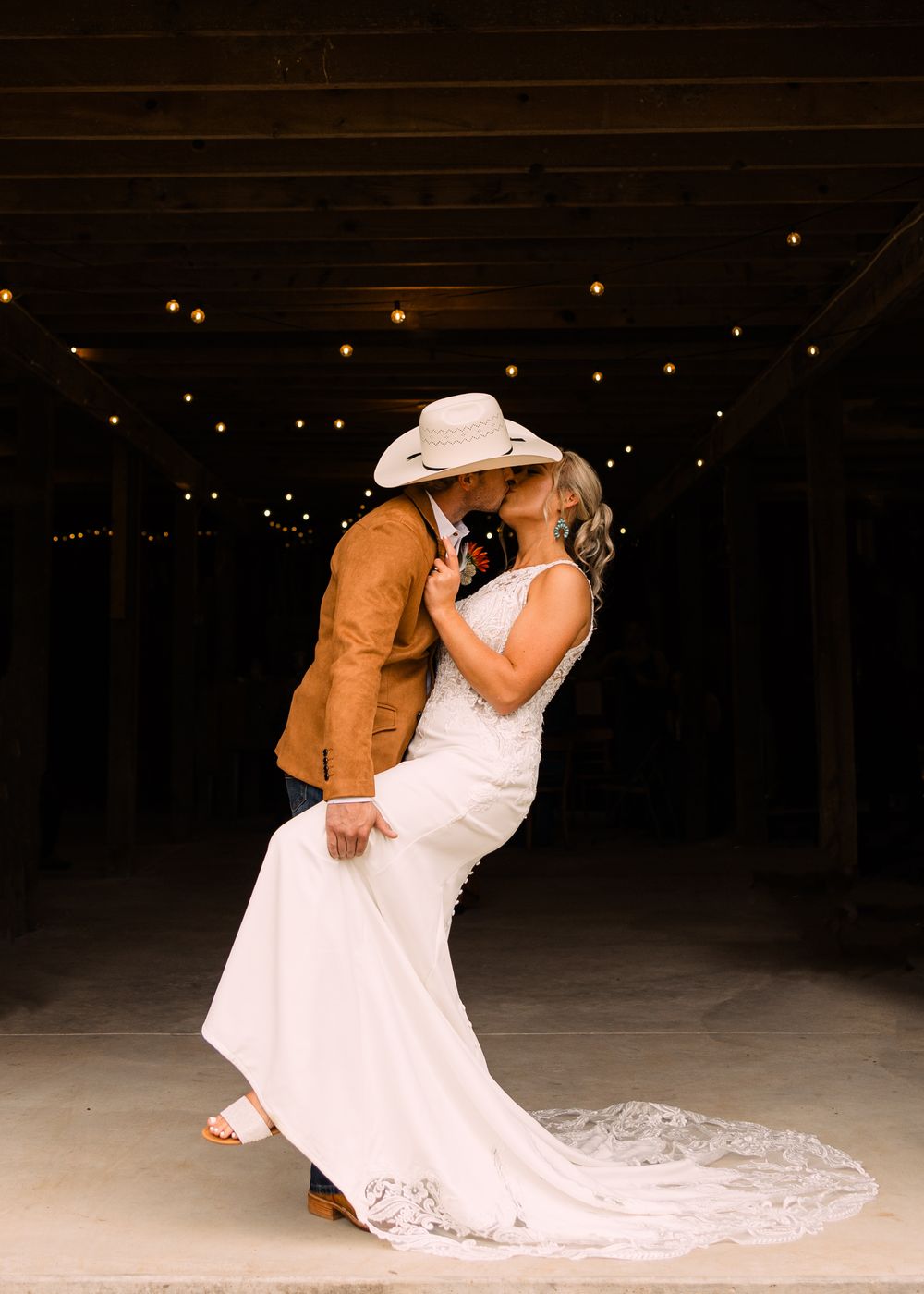 Groom in a cowboy hat dips and kisses his bride in a lace wedding dress inside a rustic barn lit by string lights.