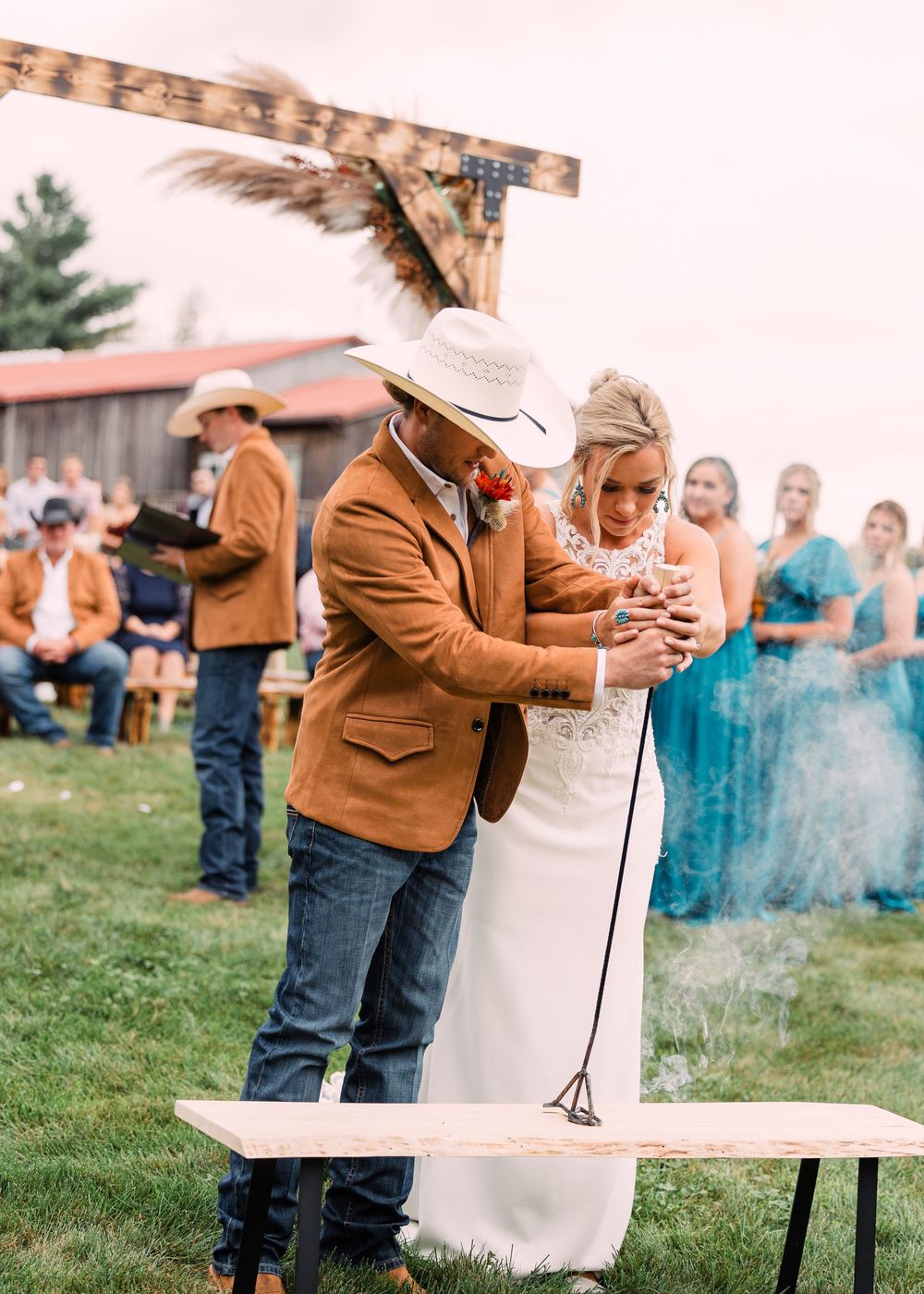 Groom in a cowboy hat and tan jacket stands with his bride during an outdoor western wedding ceremony with guests watching.