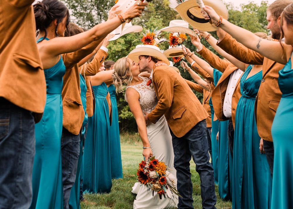 Bride and groom in cowboy hats kiss as wedding guests in teal dresses and rust jackets form an arch with raised arms.