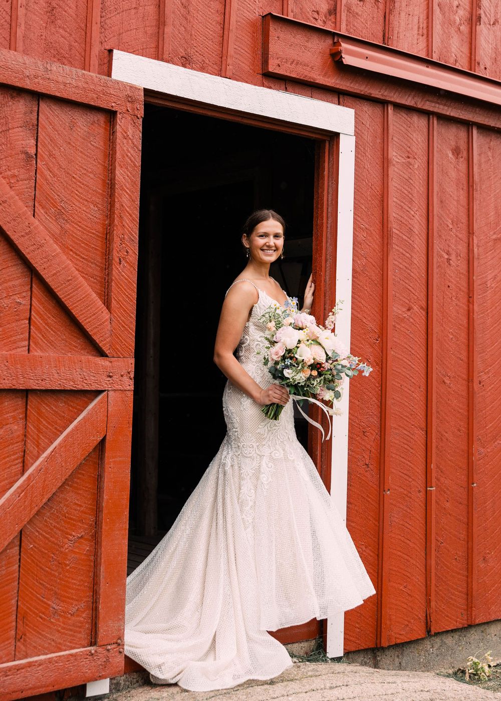 Bride in a lace mermaid wedding gown holding a pink and white bouquet stands in the open doorway of a red barn.