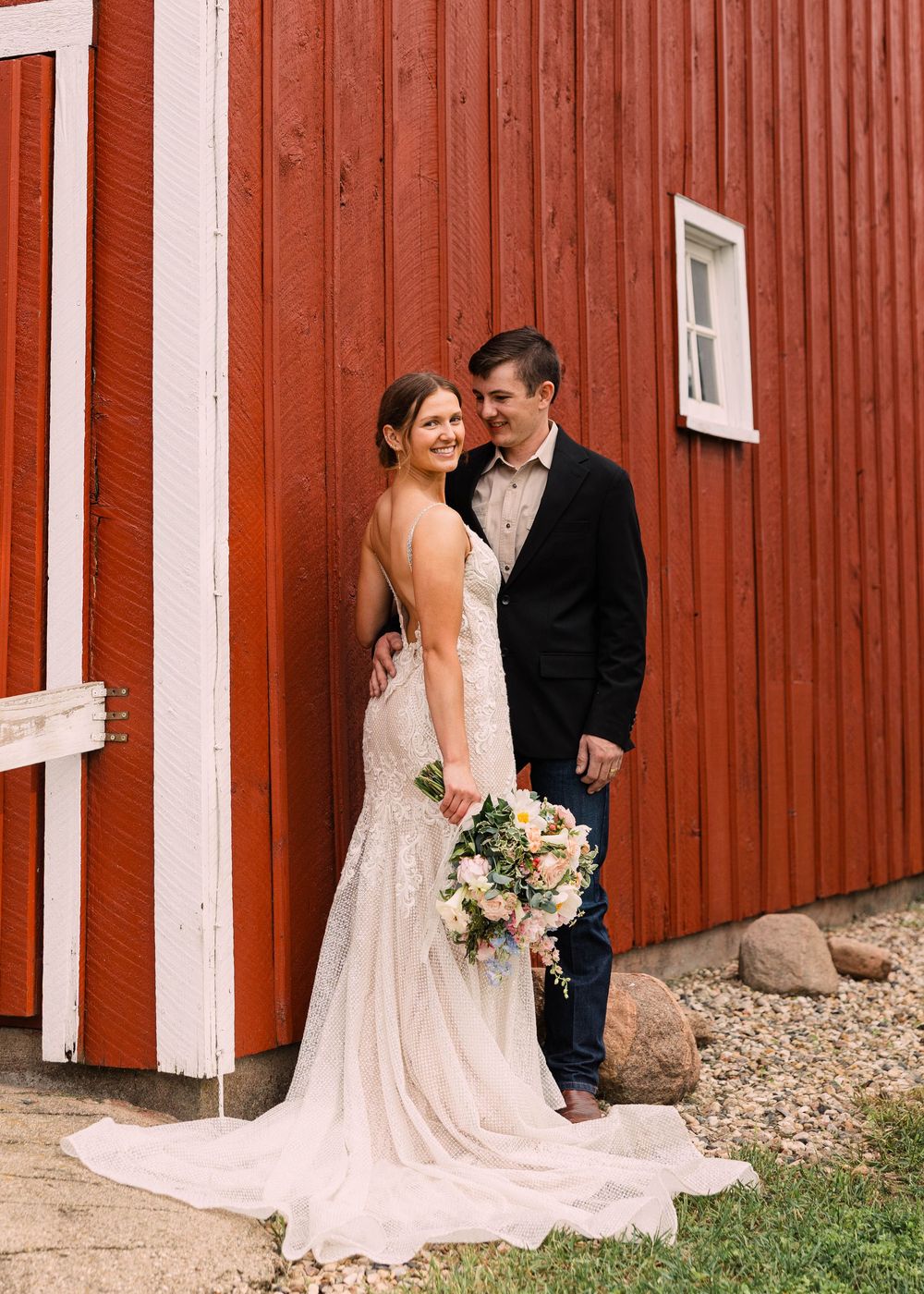 Smiling bride in a lace wedding gown holding a bouquet embraces her groom in a black jacket against a red barn wall.