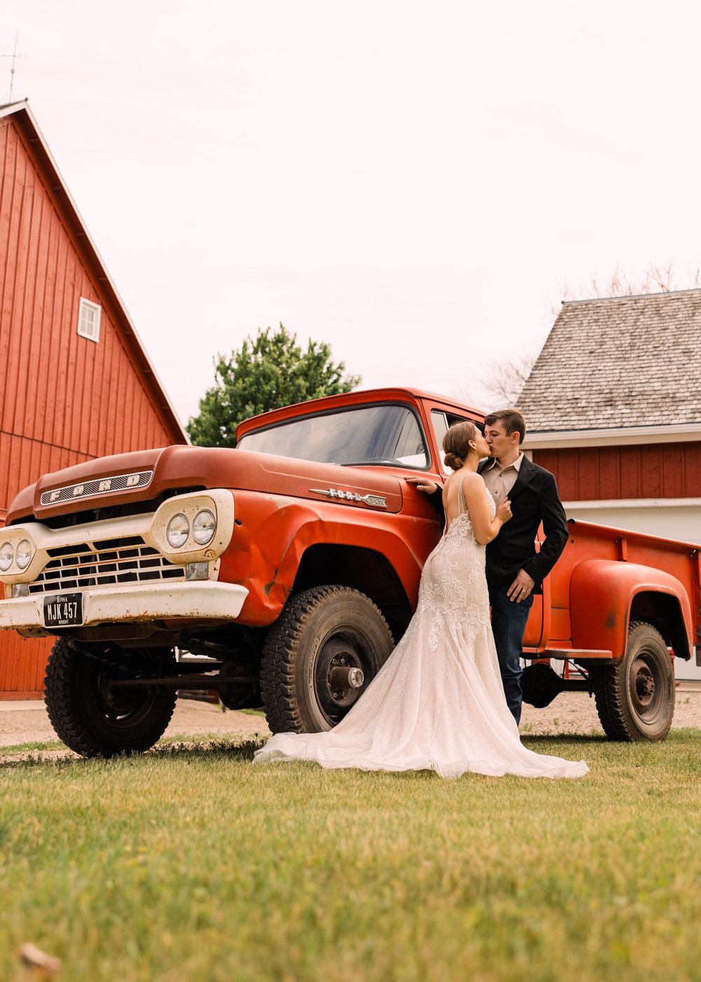 Bride and groom share a kiss beside a vintage red pickup truck in front of a red barn on a sunny grassy lawn.