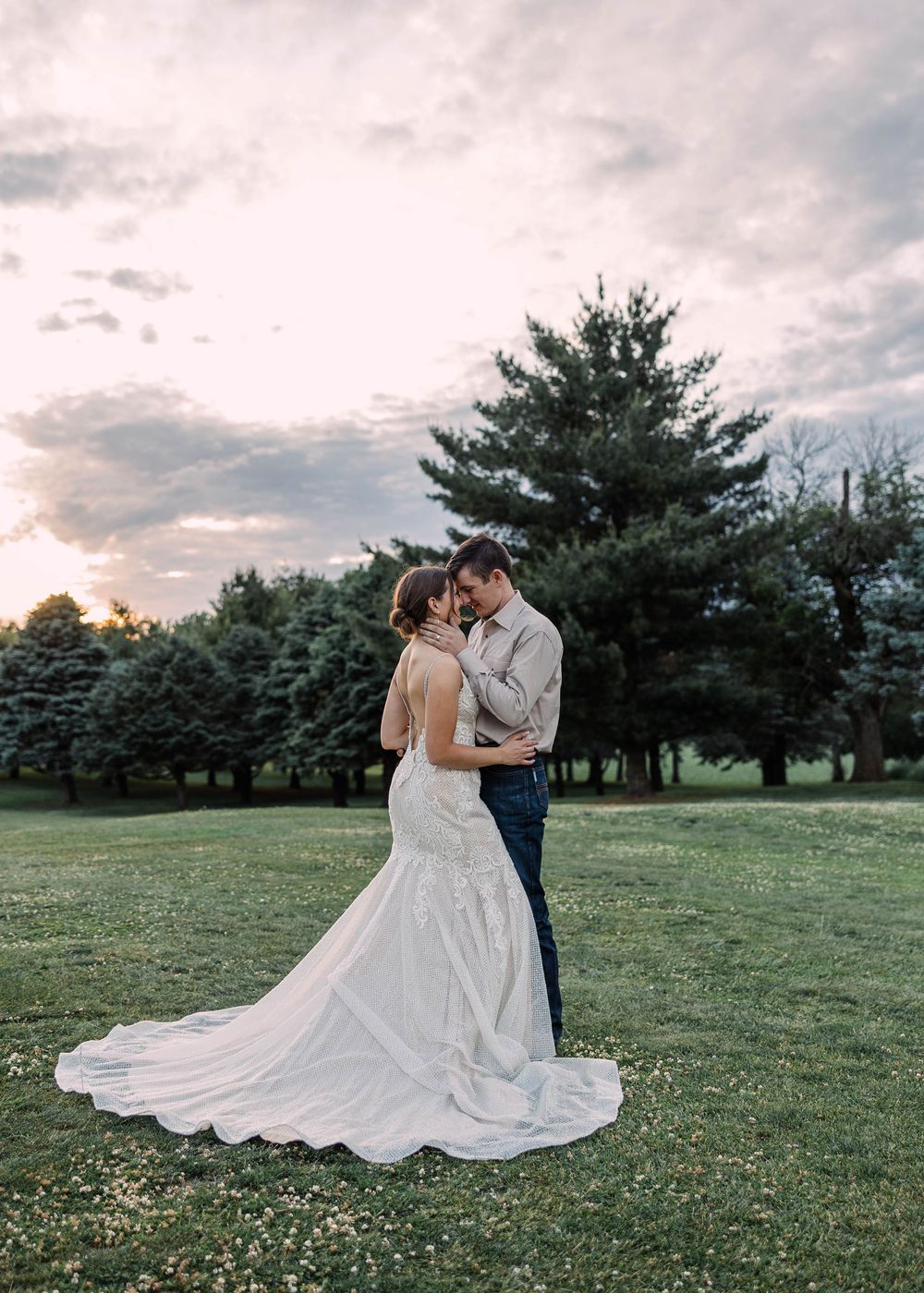 Newlyweds embrace forehead to forehead on a green lawn with evergreen trees and a soft pink sunset sky behind them.