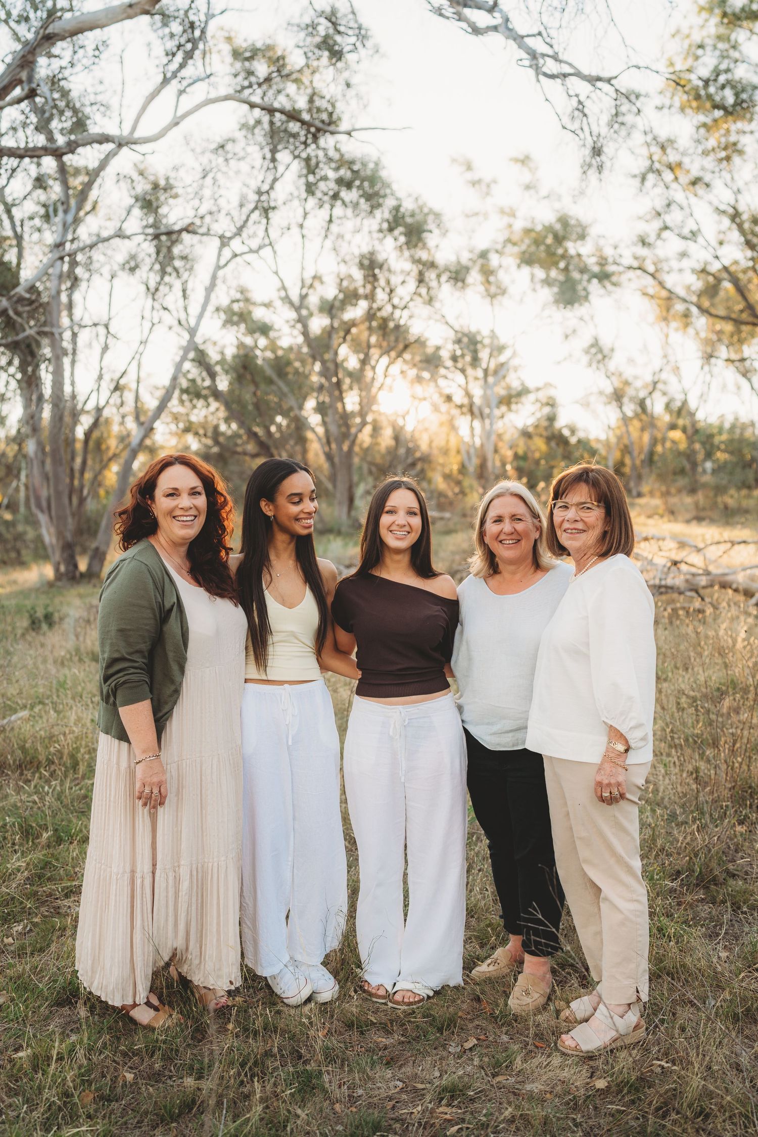 Five smiling women of different ages posing together outdoors in natural bush setting at sunset.