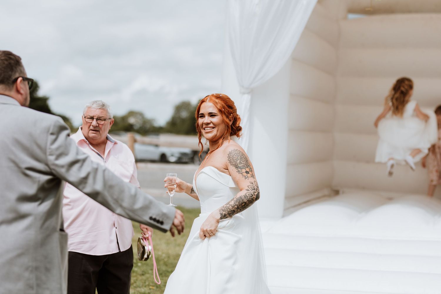 Smiling tattooed bride in white gown stands near a white bounce castle at an outdoor wedding reception.