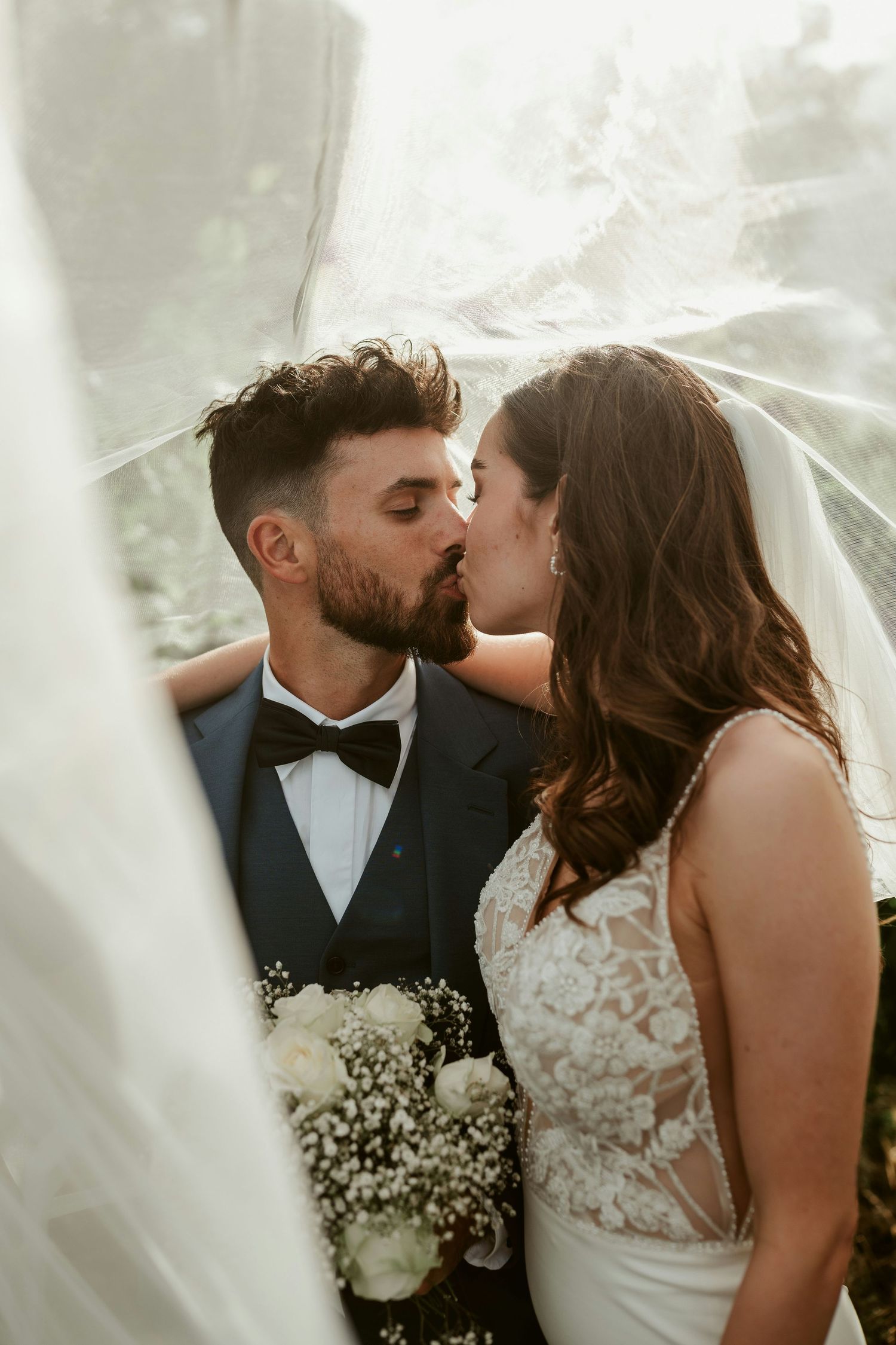 A bride and groom share a romantic kiss under a flowing veil, with the groom in a tuxedo holding white roses.