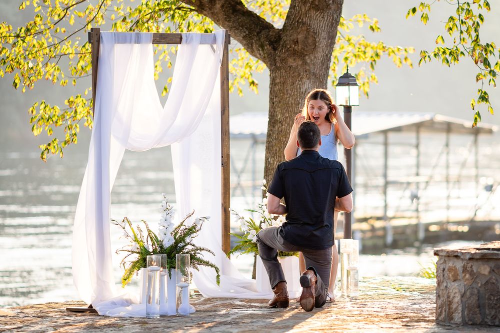 Surprise proposal by the water in Mountain Home, Arkansas, as a woman reacts beside a white draped arch under a golden-leaf tree
