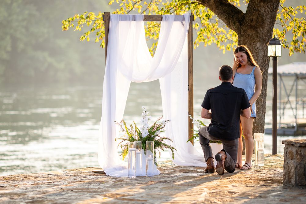 Riverside surprise proposal in Mountain Home, Arkansas, with a man kneeling by a white curtain arch and a boat dock in the background