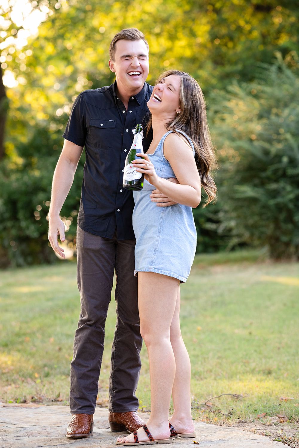 Newly engaged couple embracing with a bottle of champagne in golden evening light in Mountain Home, Arkansas
