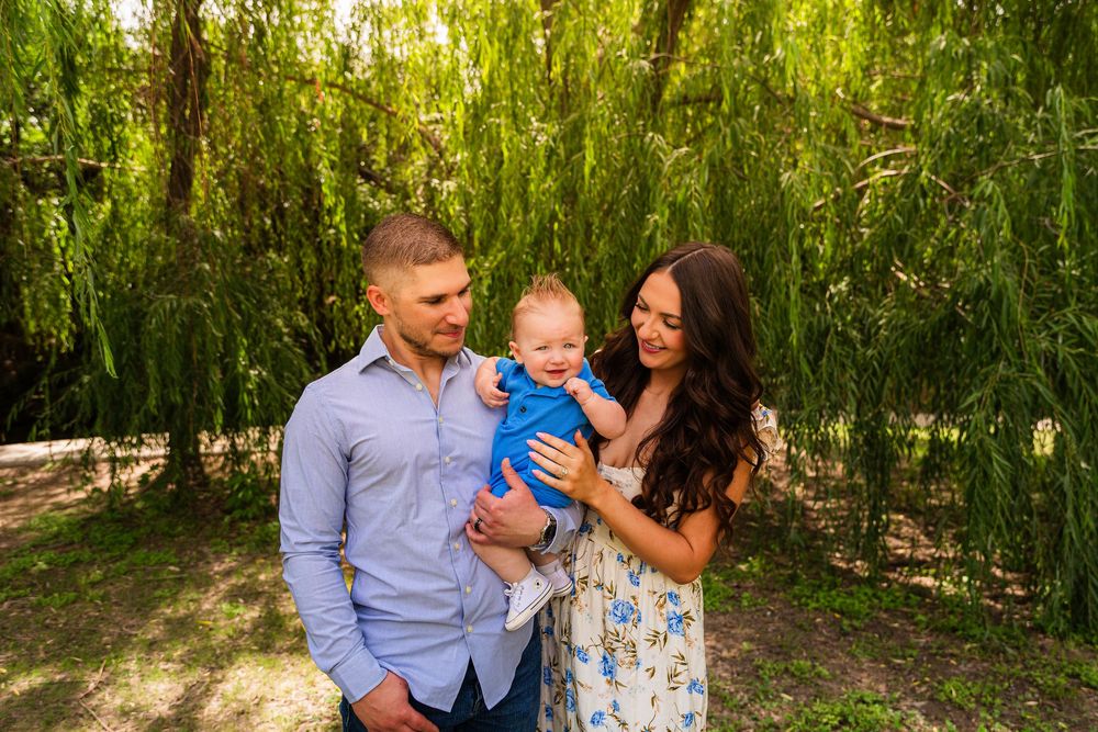 Happy young family with baby boy posing outdoors under weeping willow tree in summer.