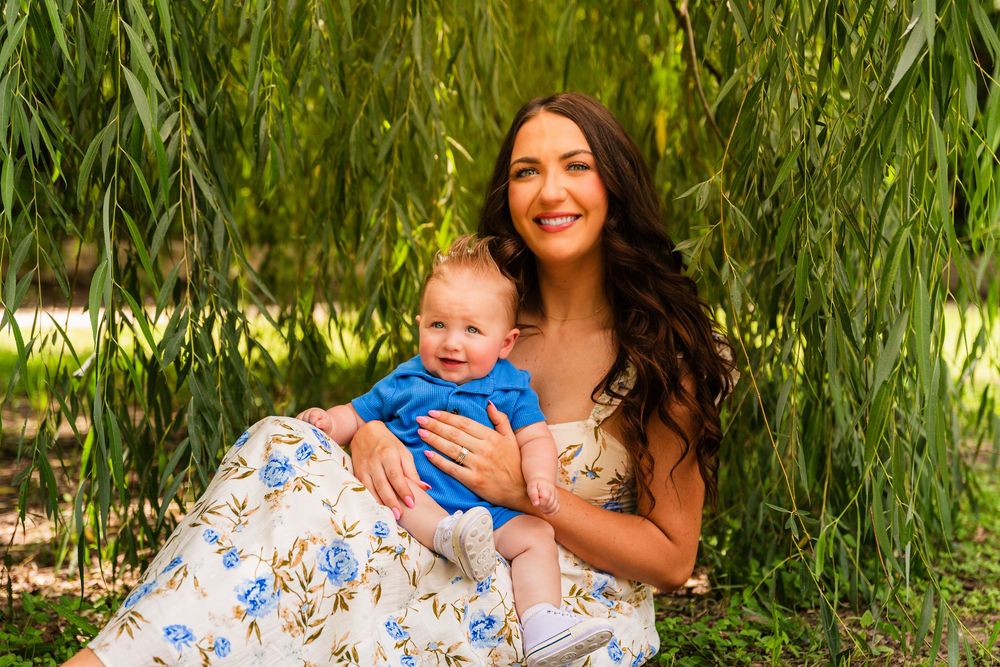 A smiling woman in a floral dress holds a baby in blue under a weeping willow tree outdoors.